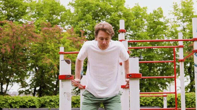Fit man doing dips on parallel bars during an outdoor calisthenics workout. Athletic male building strength through fitness training and exercise at a park. High quality 4k footage