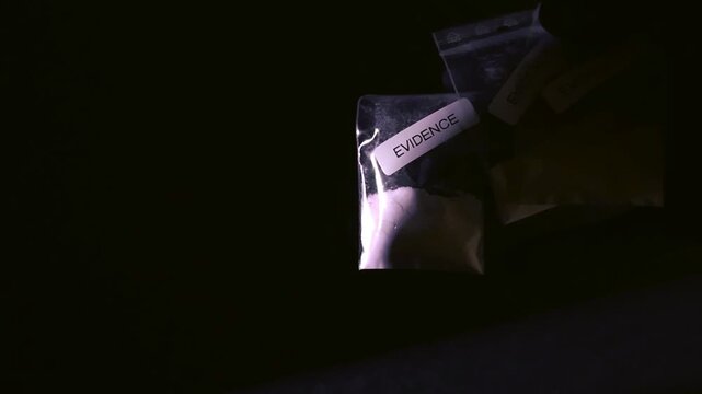 Gloved investigator's hand presenting small plastic bags filled with white powder, labeled as evidence against a dark background, suggesting a crime scene or police laboratory setting