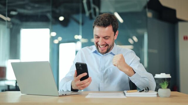 Happy businessman or IT specialist reading good news, crypto profit or casino win on mobile phone. Satisfied corporate worker, manager or freelance designer celebrating success at office desk.