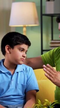Indian asian young father scolding son while sitting on sofa in modern home as upset child turns back silently, showing emotional parenting moment and family tension inside living room