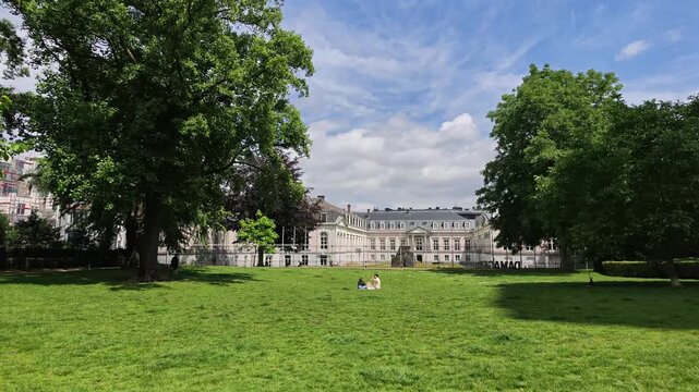 Parks of Brussels. Peaceful 4K video of a lush green lawn inside Brussels Park, with people relaxing on the grass in front of the ornate iron fence and a grand palace building in the background