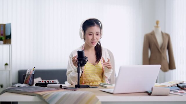 Young Asian fashion designer recording podcast episode with professional microphone and headphones in bright creative studio workspace with mannequin and laptop