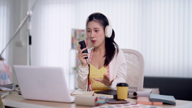 Young Asian woman podcaster recording audio show with smartphone and headphones while gesturing at laptop in bright modern home studio workspace
