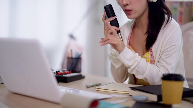 Young Asian woman podcaster recording audio content with microphone while working on laptop at bright home studio desk with coffee and notes