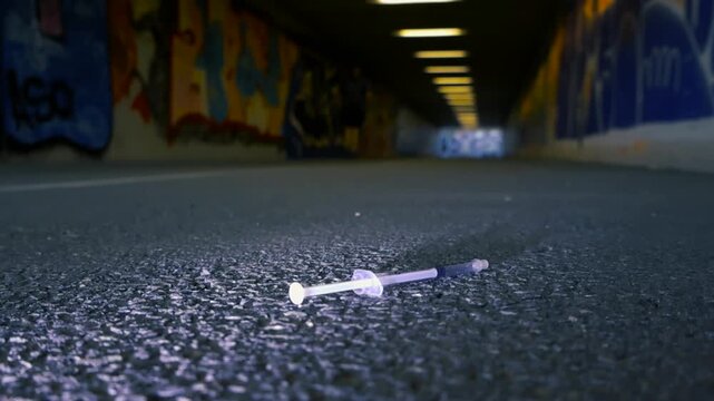 Discarded syringe on the asphalt ground of a dark city underpass with graffiti covered walls, symbolizing serious issues like drug abuse, addiction, and public health crisis