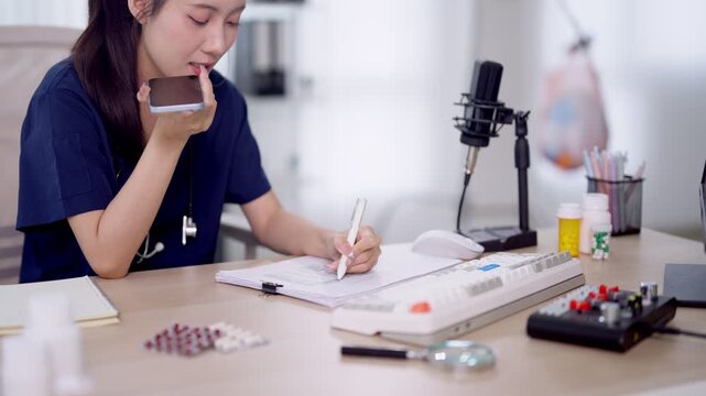 Young Asian female podcaster drinking coffee while writing notes during podcast recording session with professional microphone in bright home studio workspace