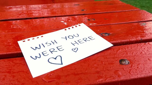 Melancholic scene with rain falling on a handwritten note left on a wet red park table expressing feelings of love and longing with the message wish you were here and two hearts