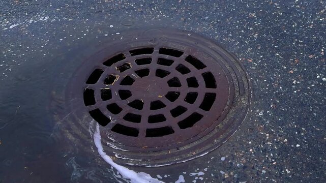 Closeup of rainwater from a heavy storm flowing down an asphalt street into a rusty cast iron manhole drain cover, managing urban runoff and preventing potential street flooding