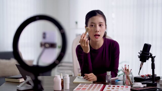 Young Asian beauty influencer demonstrating makeup foundation application while recording tutorial video with ring light and microphone in bright home studio