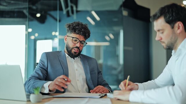 Two diverse businessmen having positive discussion at office desk with laptop and documents. Middle Eastern man in suit gesturing and talking to European colleague who listens attentively.
