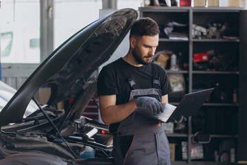 Mechanic examining car engine at auto repair shop, using laptop for vehicle diagnostic and...