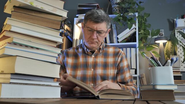 Middle aged man reading book at home office surrounded by volumes, focused self education session among literature and study materials