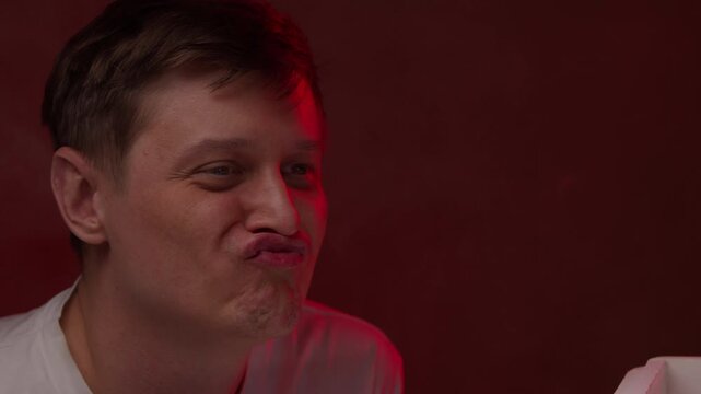 Portrait of playful caucasian man making funny faces and looking at camera with puffed cheeks and squinted eyes, portraying foolish, comic expression against dark background. Shooting in slow motion