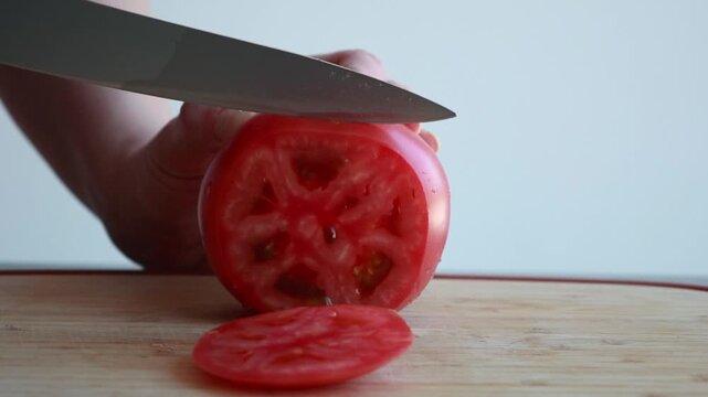 Close-up of a woman's hands slicing a large, juicy red tomato on a wooden cutting board with a sharp knife