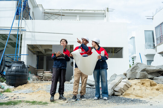 Asian construction team points at unfinished building while reviewing blueprint and laptop, planning safety inspection, quality control, project progress, site coordination meeting update plan.