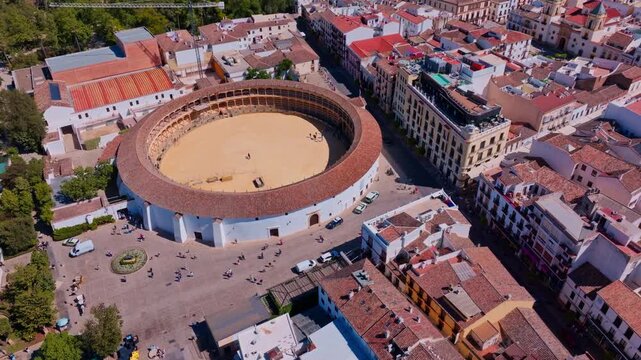 Drone footage descending forward over Ronda's bullring, framing the open sand arena against dense terracotta rooftops, street-level tourists, and ornamental plaza below.