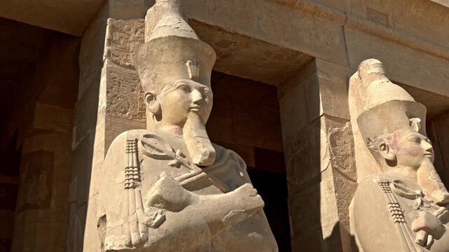 A close-up, medium shot of two monumental Osiride statues carved from limestone at the Mortuary Temple of Hatshepsut (Deir el-Bahari) in Luxor, Egypt.