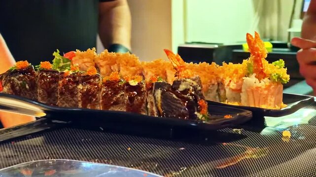 Two people observing an elaborate sushi platter with tempura shrimp rolls, teriyaki glazed pieces and tobiko on a black plate at a Japanese restaurant in Krakow, Poland.