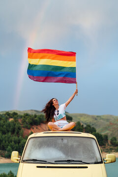 Smiling young woman holding rainbow flag sitting on van