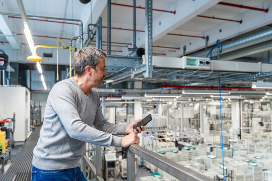 Engineer with cell phone overlooking machinery in modern production hall