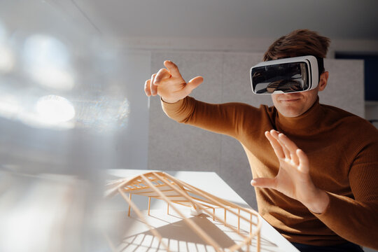 Smiling architect wearing virtual reality simulator gesturing above leaf shaped model at desk in office
