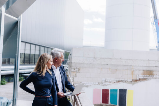 Businessman with laptop standing by colleague outside factory