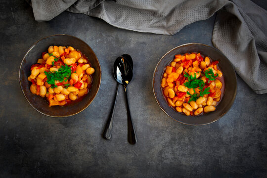 Two bowls of broad bean stew with tomatoes on black concrete