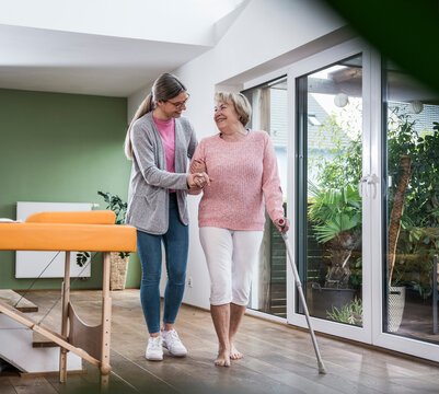 Smiling nurse helping senior woman walk with a stick indoors