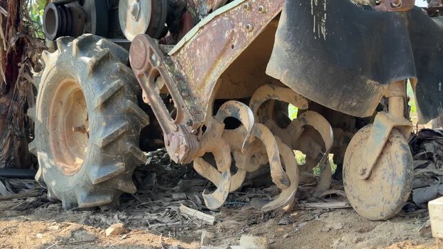Close view shows the metal wheel and rotating blade mechanism of a compact agricultural tiller placed beside a banana tree within a tropical plantation area. Agricultural equipment.