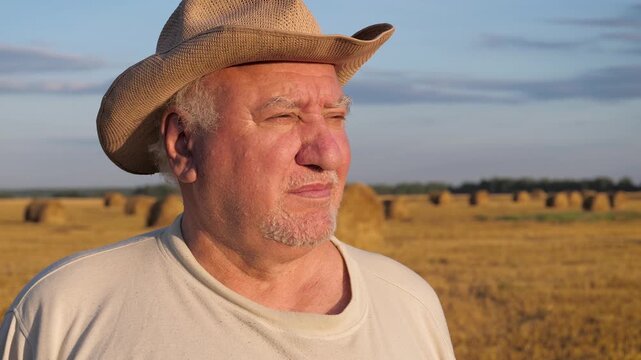 Mature male in cowboy hat standing in mown wheat field with haystacks at summer sunset. Old man contemplating successful harvest with thoughtful expression on wrinkled tanned face. Shot around farmer