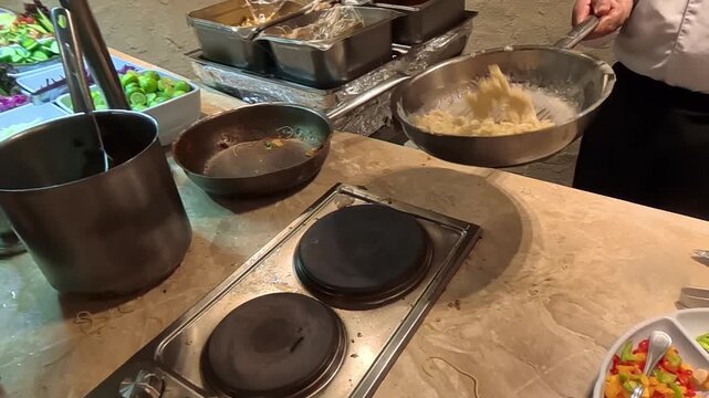 A chef stands at a marble kitchen island, mixing and presenting an array of ingredients on a long counter. Bowls of toppings, cookware on a stove, olive oil