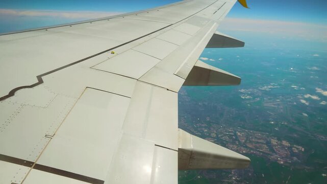 Airplane flying above cityscape with visible flaps and ailerons. Jet cruising over urban area showing detailed wing structure. Passenger aircraft gliding across skyline with view of airport and