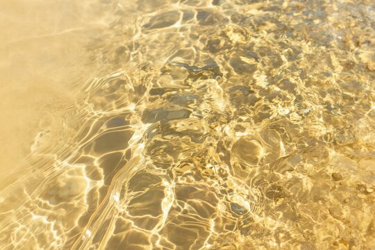 Clear yellow water ripples with bright sunlight reflections on sandy bottom. Abstract liquid texture forms wavy patterns creating serene peaceful scene perfect for summer themes.