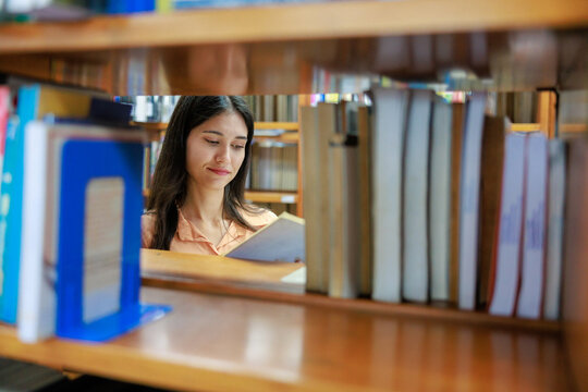 A peaceful young female student is seen through the gap of a wooden bookshelf, quietly reading and focusing on a book in a university library or local bookstore.