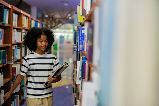 A focused young African American female student with curly natural hair stands in a bright library aisle, holding a book while carefully selecting another from a wooden bookshelf.