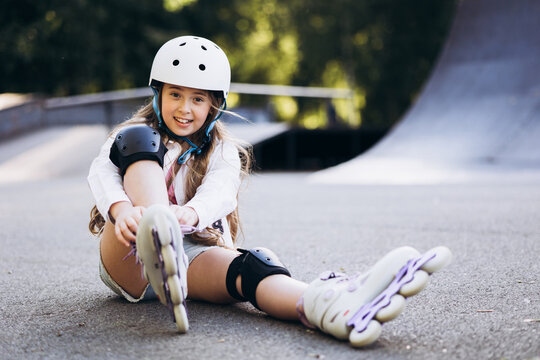 Young girl sitting on the ground, lacing rollers in a skate park, wearing a helmet and protective pads, smiling
