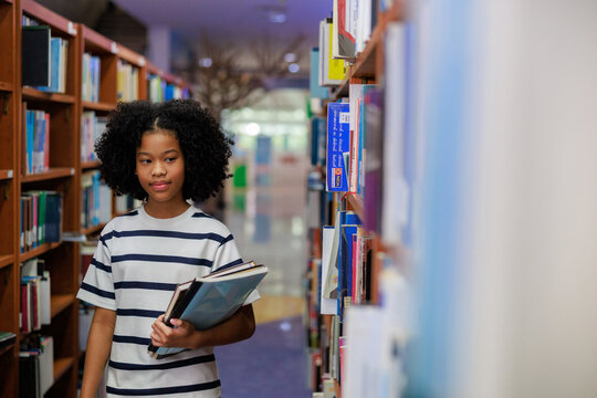 A focused young African American female student with curly natural hair stands in a bright library aisle, holding a book while carefully selecting another from a wooden bookshelf.