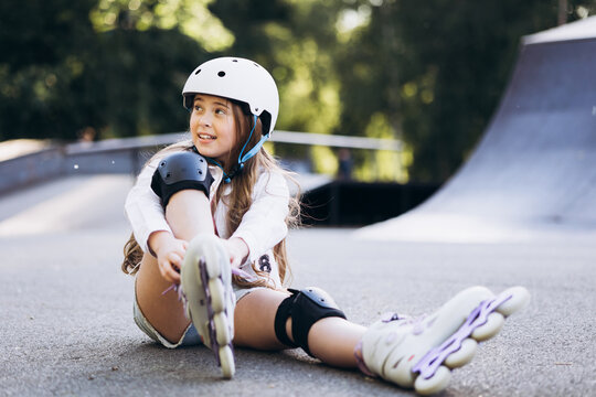 Young girl sitting on the ground, wearing a helmet and knee pads, putting on rollerblades at a skatepark