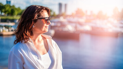 Fototapeta premium Portrait of vital brunette woman over 50 at yacht marina during golden hour, sun rays and elegant vacation lifestyle concept with copy space 