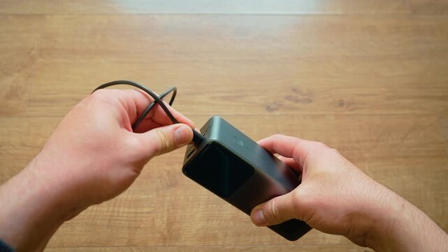Close up male hands pulling USB cable from power bank portable charger modern external battery. Disconnecting mobile charging accessory over wooden table minimal tech lifestyle indoor scene. Finishing