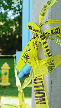 Yellow caution tape is wrapped around a pole in a park. A fire hydrant and some green space are visible in the background. The scene is well lit by sunlight in the afternoon.