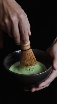 Person whisks green matcha powder with bamboo whisk in dark bowl creating foamy beverage. Traditional Japanese tea ceremony preparation, healthy drink making.