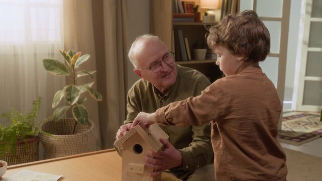 Medium shot of senior Caucasian grandfather helping young grandson assemble wooden birdhouse with screwdriver at home. Footage represents intergenerational bonding and family DIY craft activities