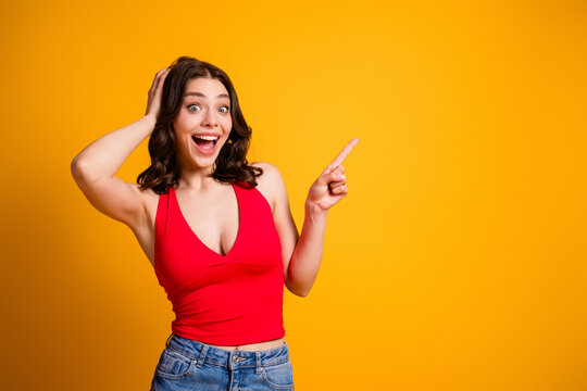 Excited young woman in red top pointing to the right with surprised, happy expression on a bright yellow background