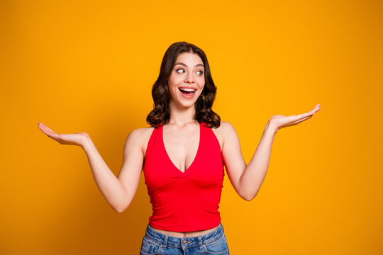 Cheerful young woman in red top presenting with palms up against vibrant orange background, smiling and looking surprised while posing in casual jeans for lifestyle and advertising use