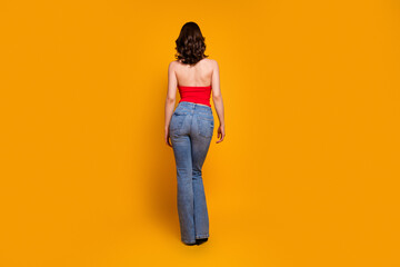 Fototapeta premium Young woman in a red tube top and blue jeans standing with her back to the camera against a bright orange background, casual fashion pose conveying confidence and minimalist studio style