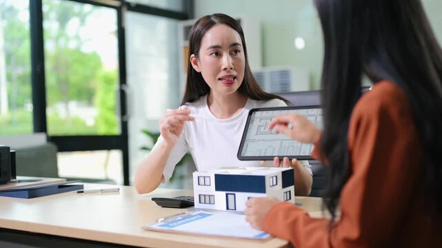 Professional architects discuss house blueprints on a digital tablet with a model home and contract on the table.
