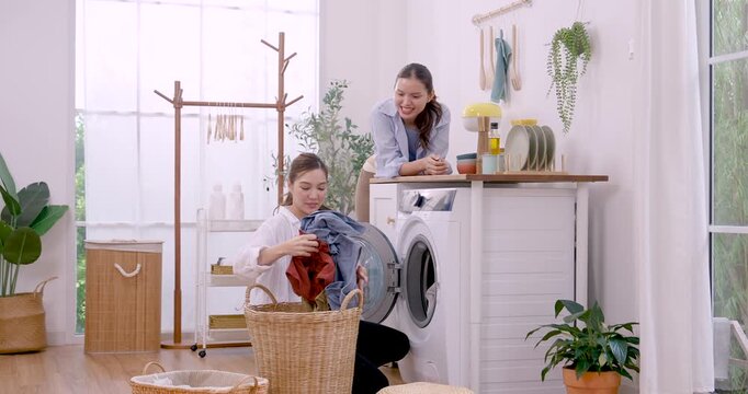 Asian LGBTQ female couple adds colorful clothing into washing machine, passing garments from basket and checking drum during cozy weekend routine, shared laundry work, support, care and love