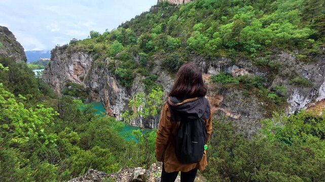 Young woman with a backpack hiking in the mountains, stopping to admire the breathtaking view of the entremon gorge and the turquoise water of the cinca river in the spanish pyrenees
