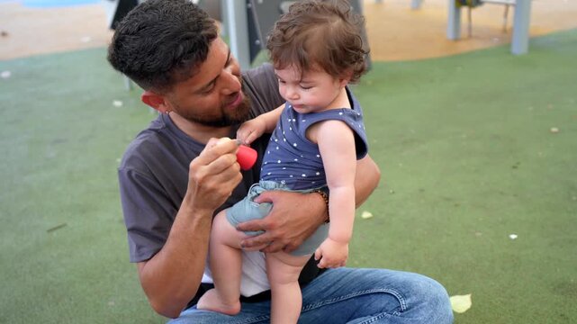 Hispanic father holding his crying toddler in his arms while trying to comfort and play with him at an outdoor playground, showing a genuine moment of parenting and paternal love